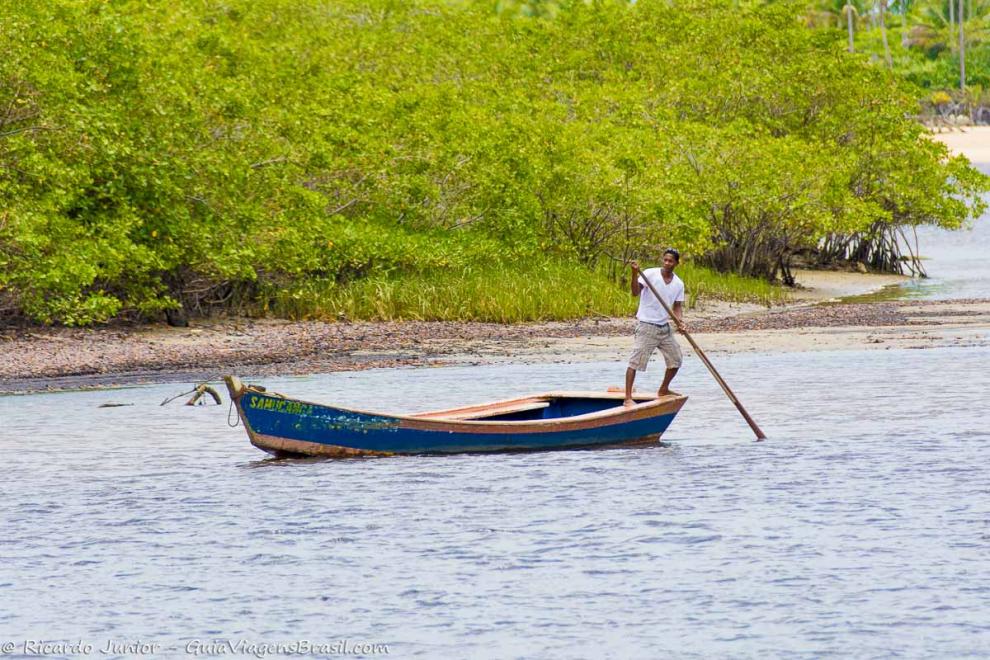 Imagem de um pescador em pé no barcos navegando nas águas calmas rio Caraiva.